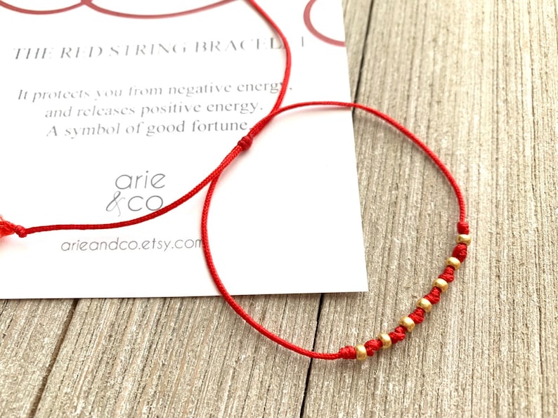 May include: Red string bracelet with gold beads. The card reads "The Red String Bracelet. It protects you from negative energy and releases positive energy. A symbol of good fortune. arie & co arieandco.etsy.com"