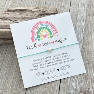 May include: A white card featuring a rainbow made from colourful paper clips and a pencil. The rainbow has a pink heart at its centre. The card reads "teach love inspire" and has a turquoise string bracelet with a silver heart charm attached. The card also includes instructions for making a wish and the text "ASK BELIEVE RECEIVE arieandco.etsy.com"