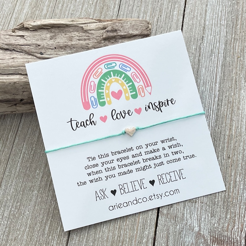 May include: A white card featuring a rainbow made from colourful paper clips and a pencil. The rainbow has a pink heart at its centre. The card reads "teach love inspire" and has a turquoise string bracelet with a silver heart charm attached. The card also includes instructions for making a wish and the text "ASK BELIEVE RECEIVE arieandco.etsy.com"