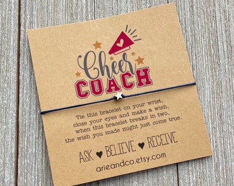 May include: A brown card with a black string bracelet and a silver star charm. The card has red and white text that says "Cheer Coach" and includes the text "Tie this bracelet on your wrist, close your eyes and make a wish, when this bracelet breaks in two, the wish you made might just come true. Ask Believe Receive arieandco.etsy.com"