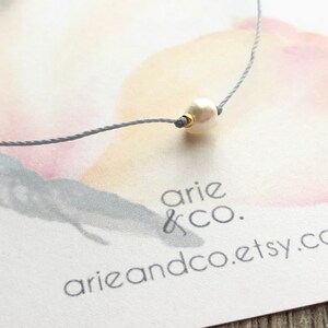 Puede incluir: Un delicado collar de cuerda gris con una sola perla blanca y un acento dorado. El collar se muestra sobre un fondo blanco con un dise&ntilde;o de mariposa de acuarela y el texto "arie & co. arieandco.etsy.com".