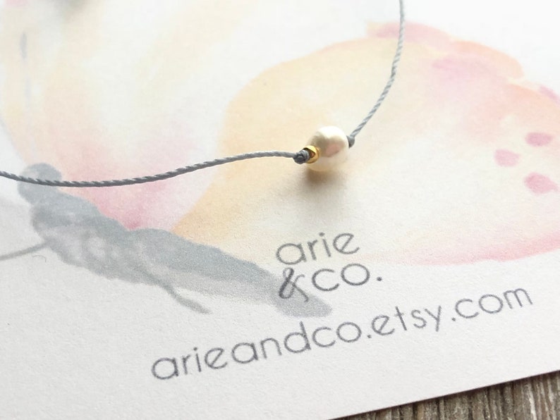 Puede incluir: Un delicado collar de cuerda gris con una sola perla blanca y un acento dorado. El collar se muestra sobre un fondo blanco con un dise&ntilde;o de mariposa de acuarela y el texto "arie & co. arieandco.etsy.com".