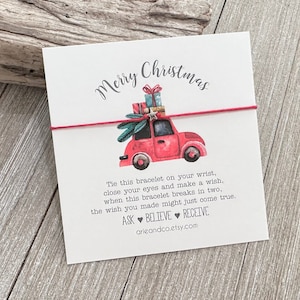 May include: Red Christmas wish bracelet with a red car carrying presents and a Christmas tree on top. The card reads "Merry Christmas" and includes the text "Tie this bracelet on your wrist, close your eyes and make a wish, when this bracelet breaks in two, the wish you made might just come true. ASK BELIEVE RECEIVE arieandco.etsy.com"