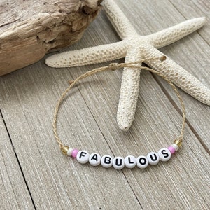 May include: A simple string bracelet with the word "FABULOUS" spelled out in white beads with black lettering. The bracelet is made with natural string and features pink and gold beads.