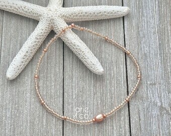 May include: A delicate anklet made with small, light brown beads and a single, larger, peach-colored pearl. The anklet is lying on a wooden surface with a white starfish.