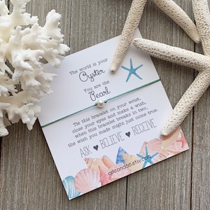 May include: A white card with a blue and green string bracelet with a pearl charm. The card has a watercolor illustration of seashells and a starfish. The text on the card reads: "The world is your Oyster You are the Pearl Tie this bracelet on your wrist, close your eyes and make a wish, when this bracelet breaks in two, the wish you made might just come true. ASK BELIEVE RECEIVE arieandco.etsy.com"