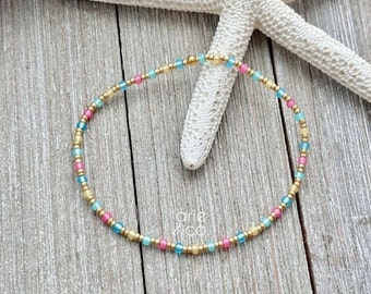 May include: A delicate gold chain bracelet with small, colourful beads in shades of pink, blue, and yellow. The bracelet is on a wooden surface with a starfish.