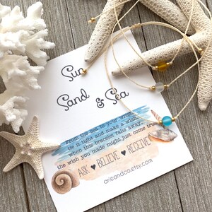 May include: A white string bracelet with a blue glass bead and gold beads. The bracelet is on a white card with the text "Sun Sand & Sea" and a message about making a wish when the bracelet falls off.