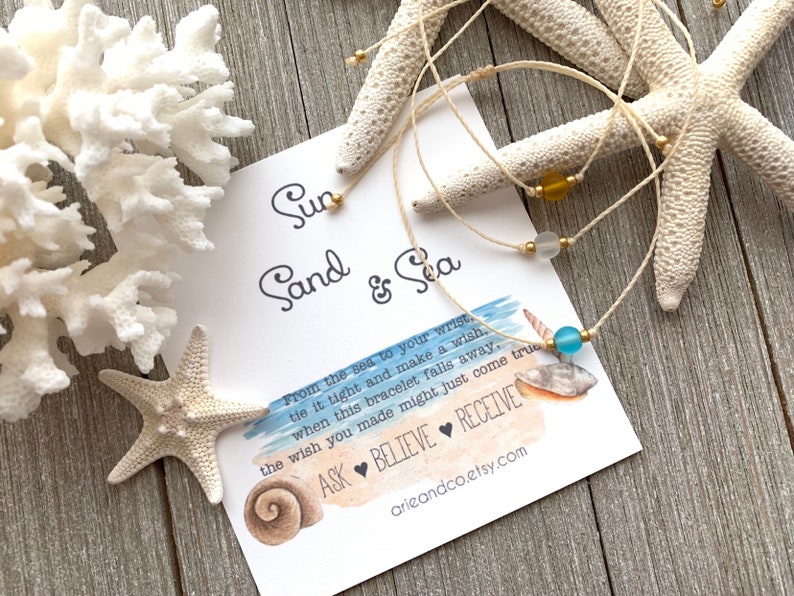May include: A white string bracelet with a blue glass bead and gold beads. The bracelet is on a white card with the text "Sun Sand & Sea" and a message about making a wish when the bracelet falls off.