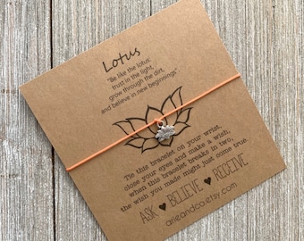 May include: A brown card with the text "Lotus" and a quote about the lotus flower. The card has a peach-colored string bracelet with a silver lotus charm attached. The text on the card reads: "Be like the lotus: trust in the light, grow through the dirt, and believe in new beginnings". The card also has the text: "Tie this bracelet on your wrist, close your eyes and make a wish, when this bracelet breaks in two, the wish you made might just come true. ASK BELIEVE RECEIVE arieandco.etsy.com"