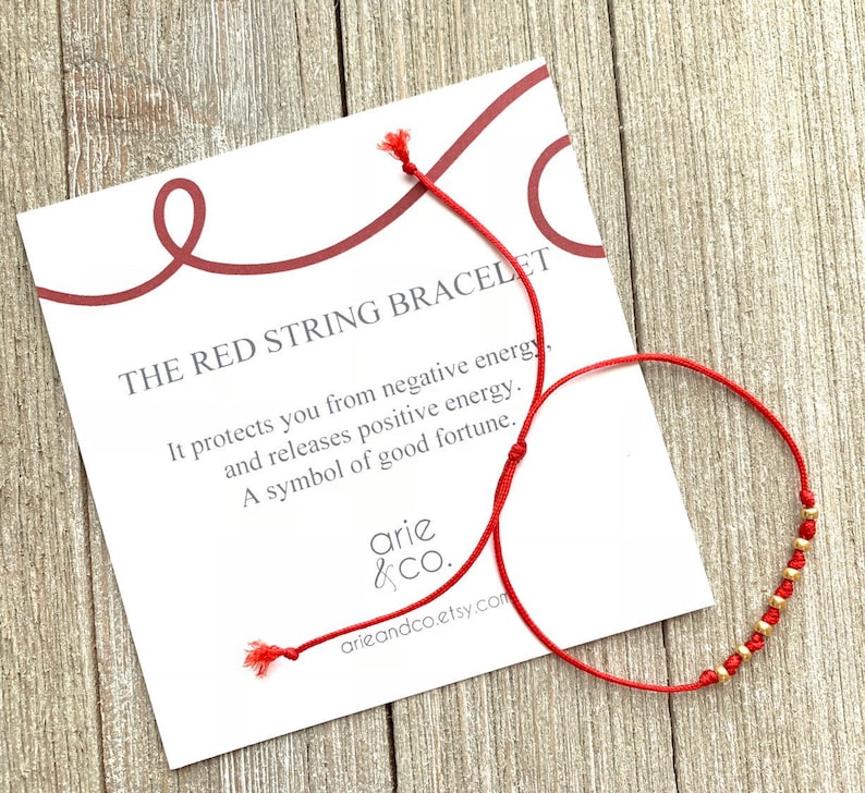 May include: Red string bracelet with gold beads. It protects you from negative energy, releases positive energy, and is a symbol of good fortune.  The text on the card reads "THE RED STRING BRACELET", "It protects you from negative energy, and releases positive energy. A symbol of good fortune."  "arie &CO. arieandco.etsy.com"