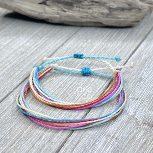 May include: Two colorful friendship bracelets made of thin, braided cords. The bracelets are layered on top of each other and feature a variety of colors, including pink, blue, white, and brown. The bracelets are tied with small blue beads.