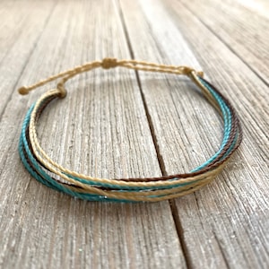 May include: A multi-strand bracelet with brown, beige, and turquoise cords. The bracelet is tied with a knot at the end.