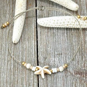 May include: A beige adjustable anklet featuring a starfish charm, white and beige beads, and gold-colored accents. The anklet is displayed on a wooden surface with starfish.