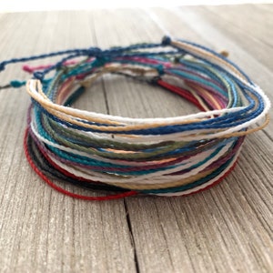 May include: A stack of colorful string bracelets in various shades of blue, green, red, white, and brown. The bracelets are made of thin, braided cord and are arranged in a circle.