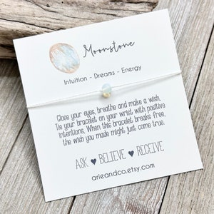 May include: White string bracelet with a moonstone bead. The card reads "Moonstone Intuition - Dreams - Energy Close your eyes, breathe and make a wish, Tie your bracelet on your wrist with positive intentions, When this bracelet breaks free, the wish you made might just come true. ASK BELIEVE RECEIVE arieandco.etsy.com"