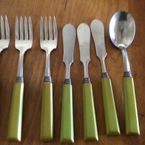 May include: A set of seven stainless steel utensils featuring solid olive green handles with a silver-toned band. The collection includes three four-tined forks, three flat-edged butter knives, and one rounded spoon, all arranged in a row.