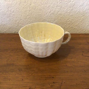 May include: A cream-colored teacup with a textured, ridged exterior and a matching handle. The interior of the cup has a pale yellow hue. The cup is sitting on a wooden surface.