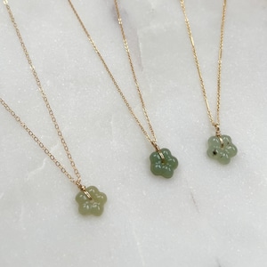 May include: Three gold chain necklaces with flower-shaped pendants. The pendants are a light green color and appear to be made of jade or a similar stone. The necklaces are displayed on a white marble surface.