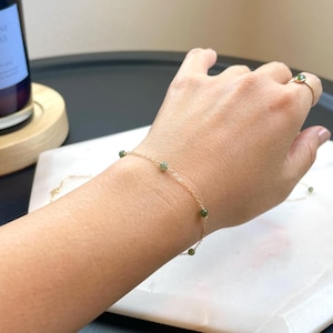 May include: A delicate gold bracelet with small, round, green gemstones evenly spaced along the chain. A matching gold ring with a single green gemstone is also visible. The jewelry is displayed on a white marble surface.