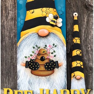 May include: A painted wooden gnome with a yellow and black striped hat, holding a beehive with flowers. The gnome is surrounded by a blue background with honeycomb patterns. The text "BEE HAPPY" is written in yellow letters below the gnome. The text "Copyright Deb Antonick" and "www.paintingwithdeb.com" are written below the text "BEE HAPPY".