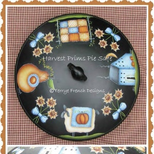 Może przedstawiać: Czarny pojemnik na ciasto z namalowanym motywem żniw. Pokrywa ma czarny uchwyt, słoneczniki, owcę z dynią i napis "Harvest Prims Pie Safe". Obręcz ma namalowane dynie i niebieskie akcenty.