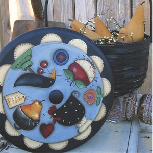 May include: A black painted wooden basket with a black metal handle. The basket is filled with pears and has a painted wooden lid with a blue background and various painted designs including a black bird, a red apple, a black chicken, and a pear. The lid has a black rim with a white scalloped edge.