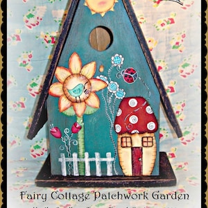 Puede incluir: Una casita de pájaros azul pintada con una escena de jardín de retazos caprichosa. La casita de pájaros presenta un girasol, una casa de setas, un sol sonriente y una mariquita. El texto "Fairy Cottage Patchwork Garden" y "Martha Smalley - Terrye French Designs - 2014" está impreso debajo de la casita de pájaros.
