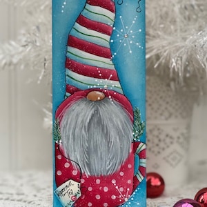 Peut inclure: Un gnome en bois peint à la main portant un chapeau rayé rouge et blanc, tenant une tasse rouge à pois blancs. La tasse a une étiquette qui dit "Peppermint Tea". L'arrière-plan est bleu avec des flocons de neige blancs.