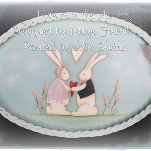 May include: A painted wooden oval sign with a white rope border. The sign features two bunnies holding a red heart. The text on the sign reads "Love is in the Air, Designed by Terrye French, Painted by Julie Shultz".
