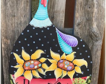 May include: A painted wooden turkey with a black and white polka dot body, a blue and white striped neck, and a bright orange hat with a white flower. The turkey is holding two yellow sunflowers with colorful centers. The bottom of the turkey is painted to look like a watermelon with the word "HOME" written on it. There is a small blue house with a red heart on the side and a green tree next to it.