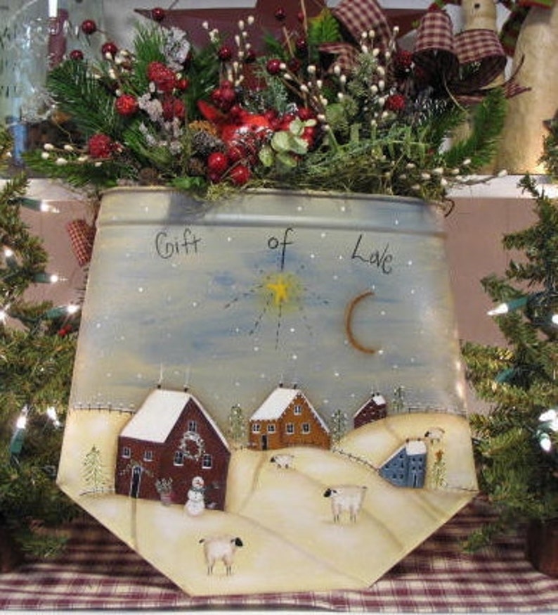 May include: A white metal container with a winter scene painted on it. The container has the words "Gift of Love" written on it. The scene includes a snowy landscape with houses, trees, and a star in the sky.