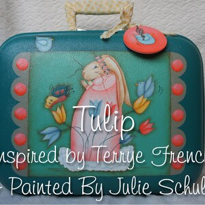 Pode incluir: Uma mala vintage azul turquesa com uma cena pintada de um coelho segurando um buquê de tulipas. A mala tem uma etiqueta rosa com um pássaro azul. O texto "Tulip" está escrito em itálico na mala. O texto "Inspired by Terrye French & Painted by Julie Schultz" está escrito abaixo da imagem.