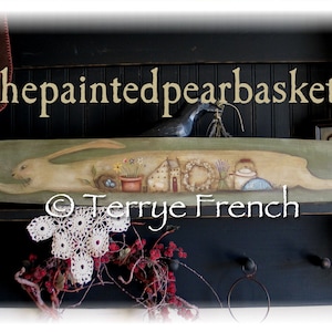 May include: A painted wooden breadboard with a green background and a white rabbit running across it. The board also features a small house, a wreath, a pot of flowers, and a bird. The text "thepaintedpearbasket" and "© Terrye French" are visible on the board.