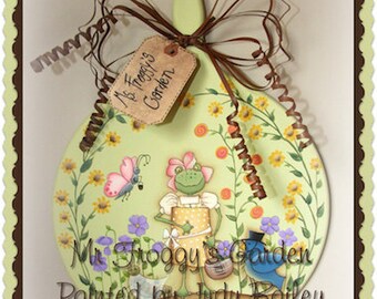 May include: A painted wooden cutting board with a green background and a frog wearing a hat and holding a watering can. The board is decorated with flowers and the text "Mr. Froggy's Garden Painted by Judy Bailey Inspired by Terrye French".