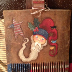 May include: A wooden comb painted with a patriotic theme. The comb has red, white, and blue stripes. A whimsical painted Santa Claus with a white beard and a red hat is on the comb. The comb is decorated with a star and holly.