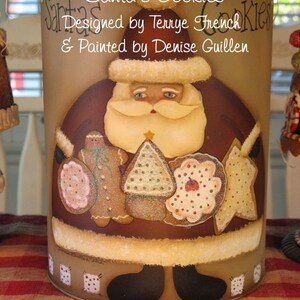 Può includere: Un barattolo per biscotti marrone con un'immagine dipinta di Babbo Natale che tiene dei biscotti. Il barattolo ha il testo "Santa's Cookies" e "Designed by Terrye French & Painted by Denise Guillen".