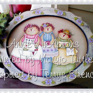 Könnte beinhalten: Ein runder Holztablett mit einer bemalten Szene von drei Frauen in Schürzen. Das Tablett ist mit einem blauen und weißen Rand und lila Blumen verziert. Der Text "Annie's in Aprons Painted by Maryjo Tuttle, Inspired by Terlye French" ist auf dem Tablett geschrieben.