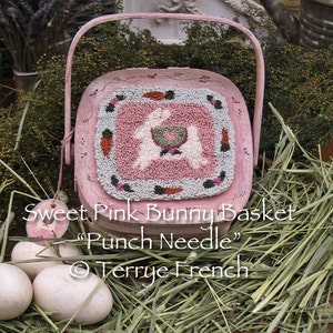 May include: A pink wooden basket with a handle, decorated with a floral design. The basket has a square punch needle embroidery of two white bunnies with a green and pink heart in the center. The text "Sweet Pink Bunny Basket "Punch Needle" © Terrye French" is written below the basket.