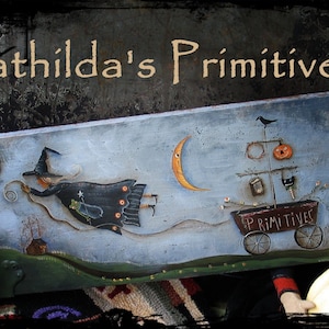 May include: A painted wooden sign depicting a whimsical scene of a witch flying on a broomstick with a crescent moon in the background. The sign also features a small wagon with the words "Primatives" painted on the side. The sign is decorated with a black crow, a pumpkin, and other details.