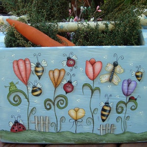 May include: A blue wooden box with a painted scene of a whimsical garden. The box features flowers, bees, butterflies, and ladybugs. The flowers are painted in shades of pink, orange, yellow, and purple. The bees are yellow and black, and the butterflies are yellow and white. The ladybugs are red with black spots.