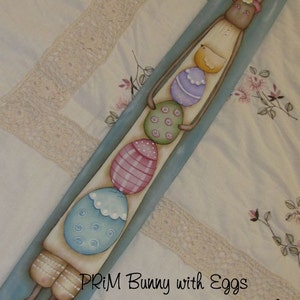 May include: A painted wooden bunny holding a stack of colorful Easter eggs. The bunny is wearing a pink flower headband and has a light blue background. The text "PRIM Bunny with Eggs" is written below the bunny, followed by "Original design by Terrye French" and "Painted by Deb Antonick".