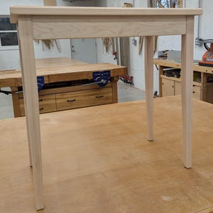 May include: A simple, unfinished wooden end table with a rectangular top and four legs. The table is made of light-colored wood and has a natural finish.