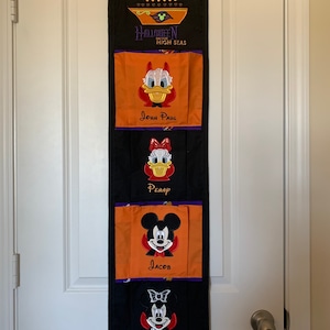 May include: A black and orange Halloween-themed fabric wall organizer with four pockets. The top pocket has the text "The Cooper Family" and "Halloween on the High Seas". The other pockets have embroidered images of Mickey Mouse and Donald Duck characters and the names "John Paul", "Penny", "Jacob", and "Priscilla".