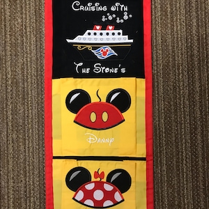 Peut inclure: Un organisateur en tissu jaune avec deux poches, chacune représentant une tête de Mickey Mouse ou de Minnie Mouse avec un nom en dessous. Le haut de l'organisateur est composé d'un panneau en tissu noir avec le texte "Cruising with The Stone's" et une silhouette blanche d'un navire de croisière.