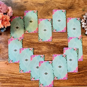 May include: A heart-shaped arrangement of ten teal-colored cards with pink floral accents and white designs. The cards are laid out on a wooden surface.