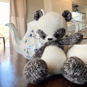 May include: A white and gray knitted panda bear toy with black eyes and a black nose. The panda is sitting on a wooden table with a white teapot and a white bowl in the background.
