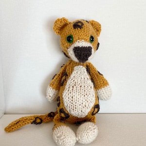 May include: A knitted cheetah toy with brown spots, white fur, and green eyes. The toy is sitting on a white surface.