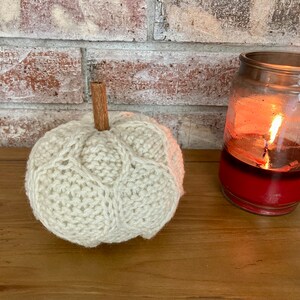 May include: A white knitted pumpkin with a cinnamon stick stem. The pumpkin is sitting on a wooden surface next to a lit candle in a glass jar.