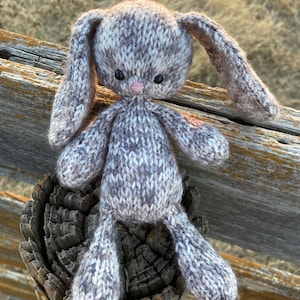 May include: A knitted gray and white bunny rabbit with pink nose and black eyes sitting on a wooden fence post.